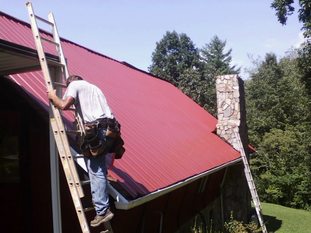 Roofer on a ladder beside a red metal roof and stone chimney — Soffit & Fascia Repair by Roofing Brothers in Tracy City